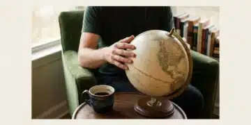 A person tracing lines on a vintage globe, symbolizing the interconnectedness of ideas, culture, and future in modern living, with colorful books in the background.